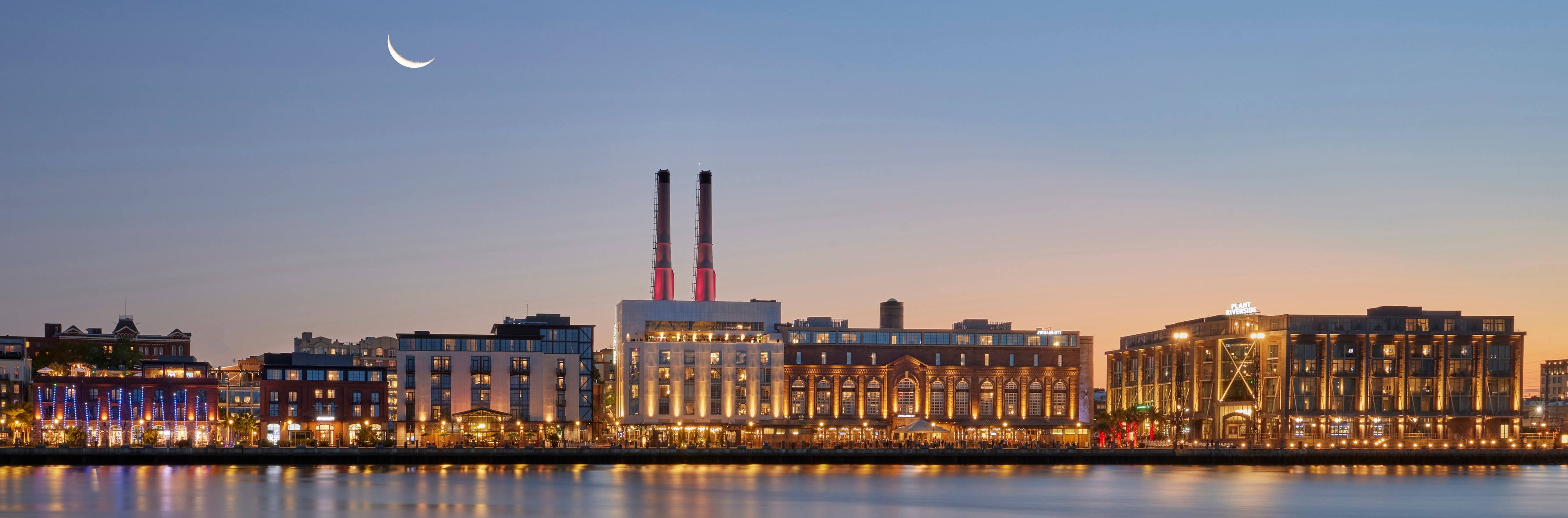 Nighttime view of River Street across the Savannah River — illuminated historic riverfront warehouses reflecting on the water